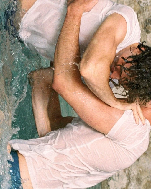 Two men in wet white t-shirts embrace in water with a stone background, creating an intimate and natural scene.