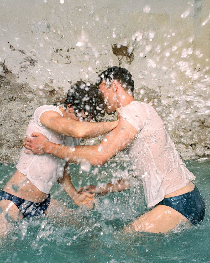Two adult men, both fair-skinned, embrace in shallow water, wearing wet white t-shirts and navy swim briefs, with splashes of water around them.
