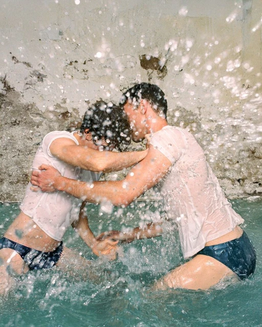 Two adult men, both fair-skinned, embrace in shallow water, wearing wet white t-shirts and navy swim briefs, with splashes of water around them.