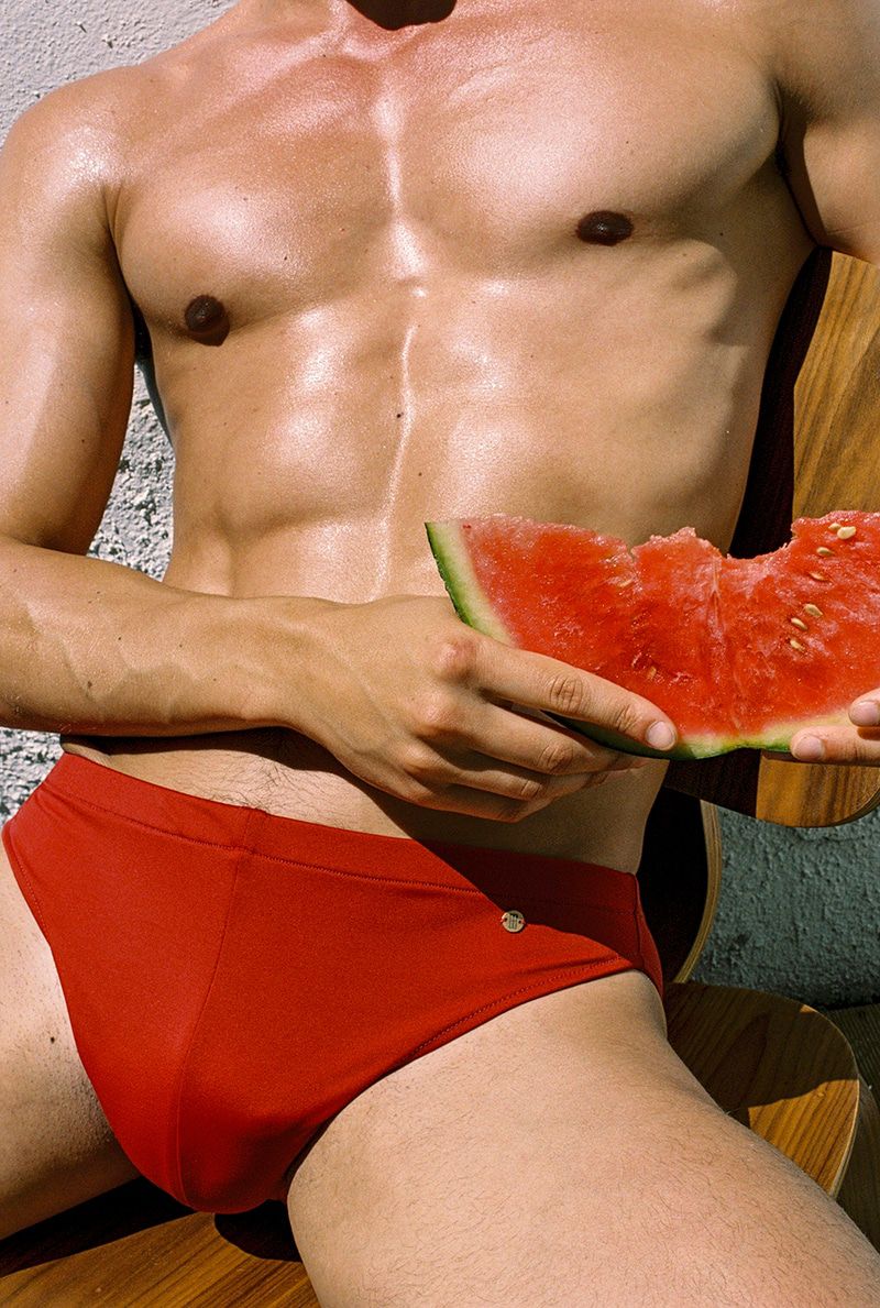 Torso shot of an adult male with a slice of watermelon while seated. He is wearing red swimwear with a bright background.