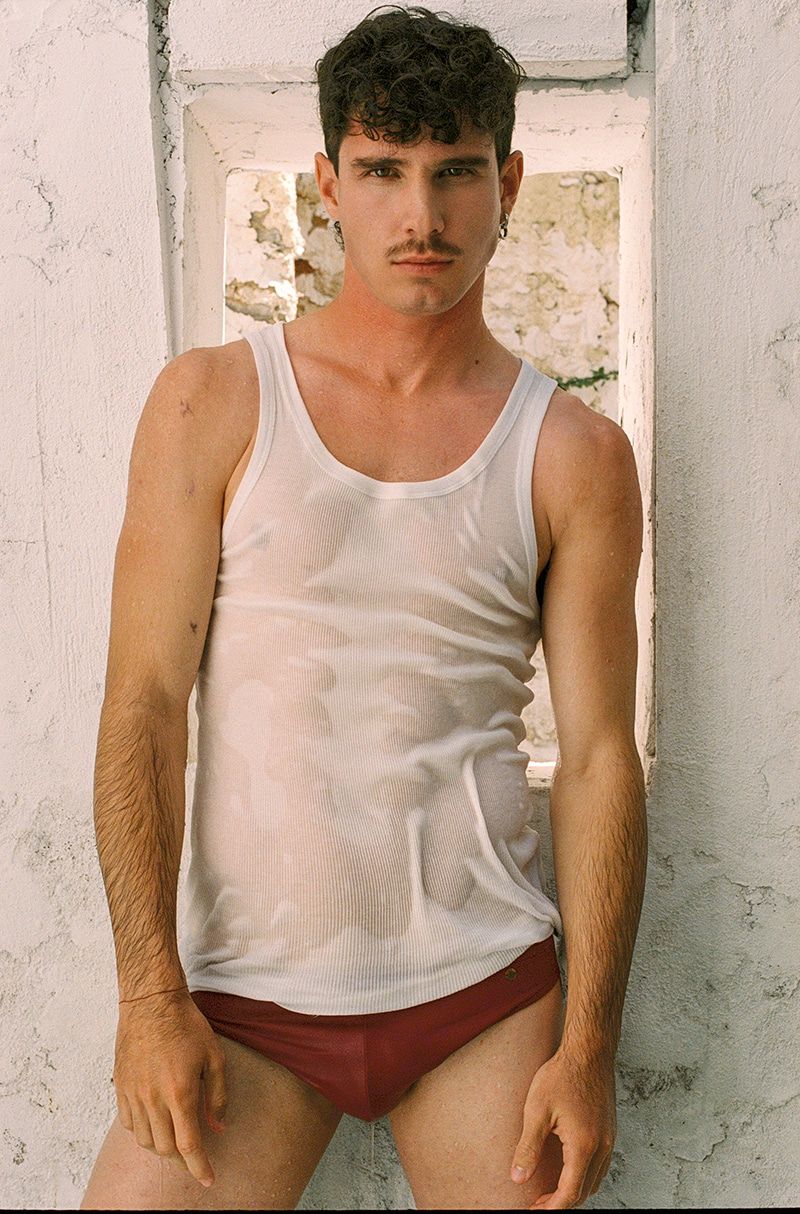 An athletic adult male poses indoors wearing a white tank top and maroon briefs next to a weathered white wall with a window opening.