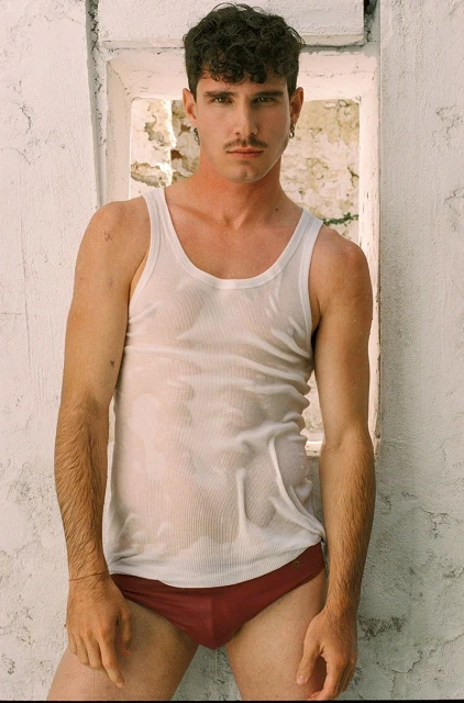 An athletic adult male poses indoors wearing a white tank top and maroon briefs next to a weathered white wall with a window opening.