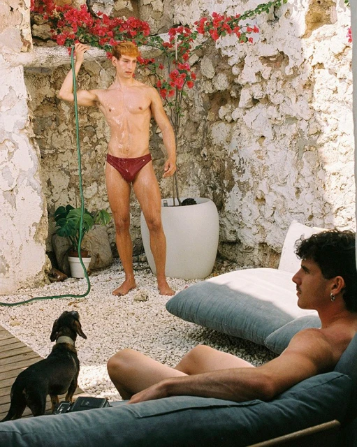 Two men in swimwear outdoors. One is standing with a hose, while the other is reclining on a cushioned lounge chair outdoors with a small dog.