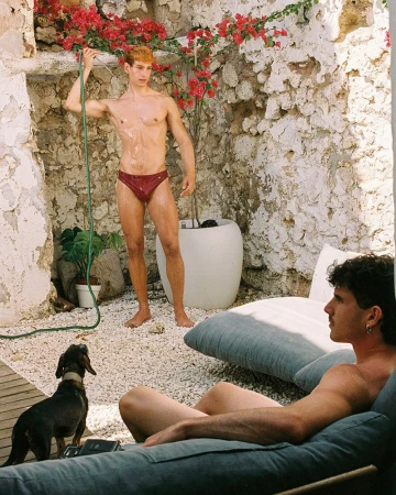 Two men in swimwear outdoors. One is standing with a hose, while the other is reclining on a cushioned lounge chair outdoors with a small dog.