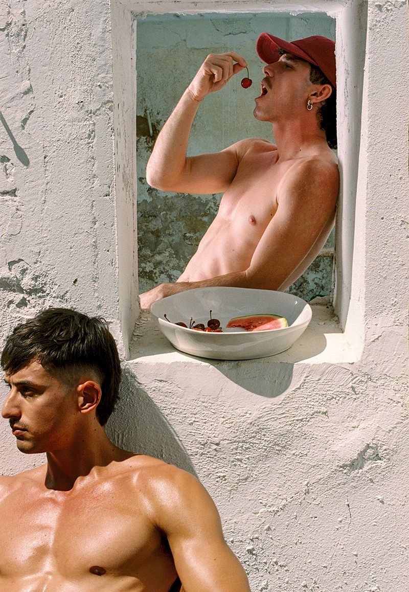 Two shirtless men in an outdoor setting; one eating a cherry in a window frame, the other resting below in natural sunlight.