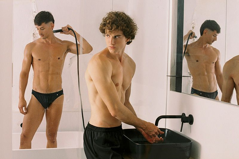 Two adult males pose in a bright, minimalist bathroom setting. One stands in a tub holding a showerhead, while the other washes his hands.