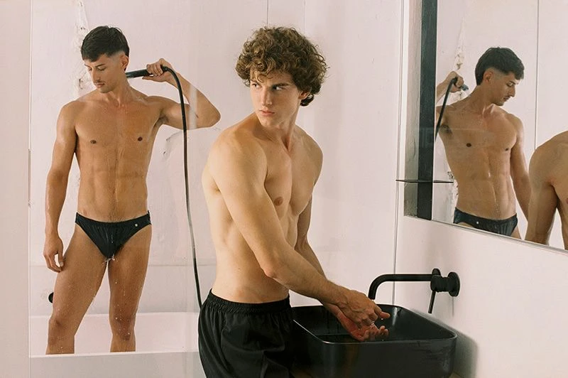 Two adult males pose in a bright, minimalist bathroom setting. One stands in a tub holding a showerhead, while the other washes his hands.