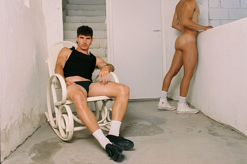 Two adult men in an indoor setting. One man poses sitting on a white rocking chair, while the other stands against a white wall.