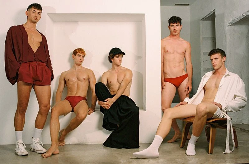 Five men in an indoor setting, featuring two shirtless men in swimwear, a man in a casual top and shorts, and two men in robes or loose shirts.