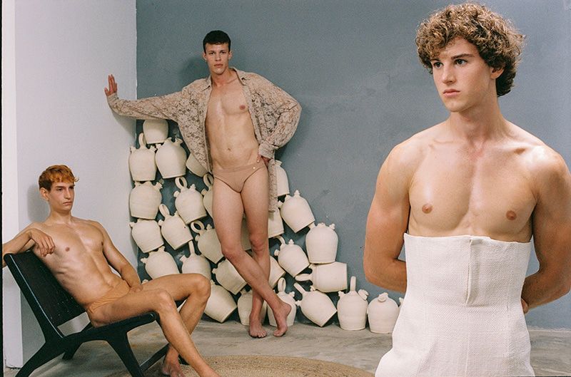 Three adult males pose indoors. One sits in a black chair, another stands with white pottery behind him, and one stands with a white cloth around his torso.