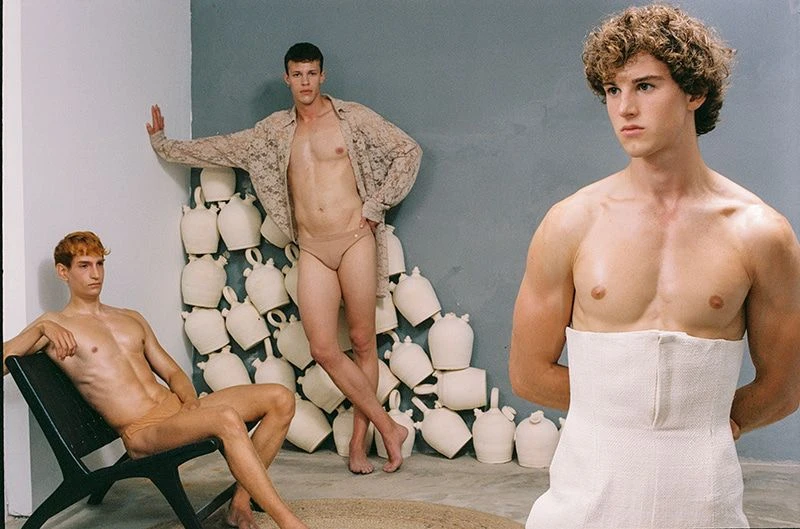 Three adult males pose indoors. One sits in a black chair, another stands with white pottery behind him, and one stands with a white cloth around his torso.