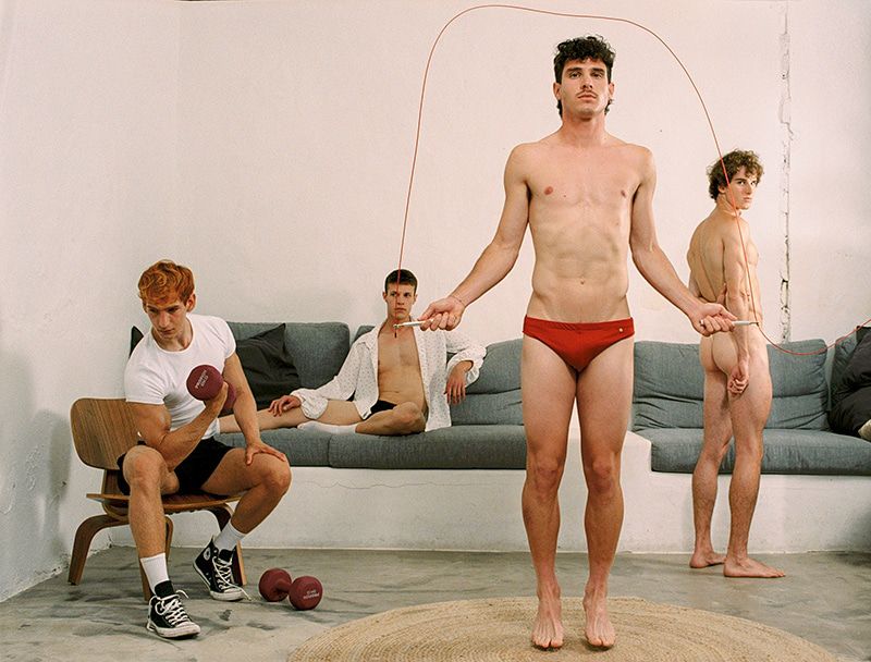 Four young adult men pose in an indoor setting; one jumps rope, another lifts a dumbbell, a third lounges on a couch.
