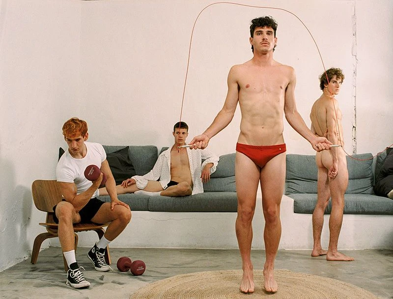 Four young adult men pose in an indoor setting; one jumps rope, another lifts a dumbbell, a third lounges on a couch.