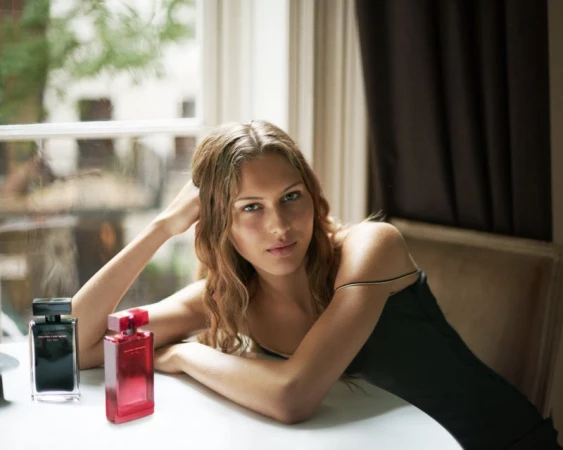 Indoor shot of a young woman with wavy blonde hair posing next to two perfume bottles in front of a window, illuminated by soft, natural light.