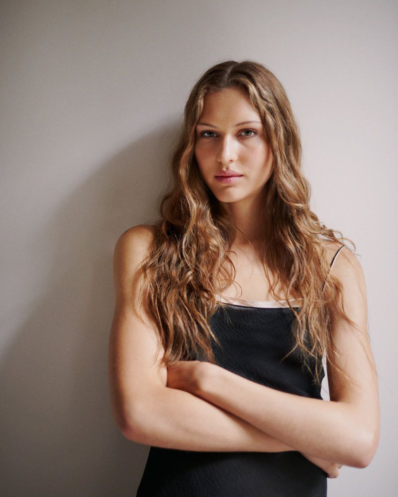 Portrait of a young adult woman with long brown wavy hair wearing a black tank top, arms crossed, in front of a light gray wall.