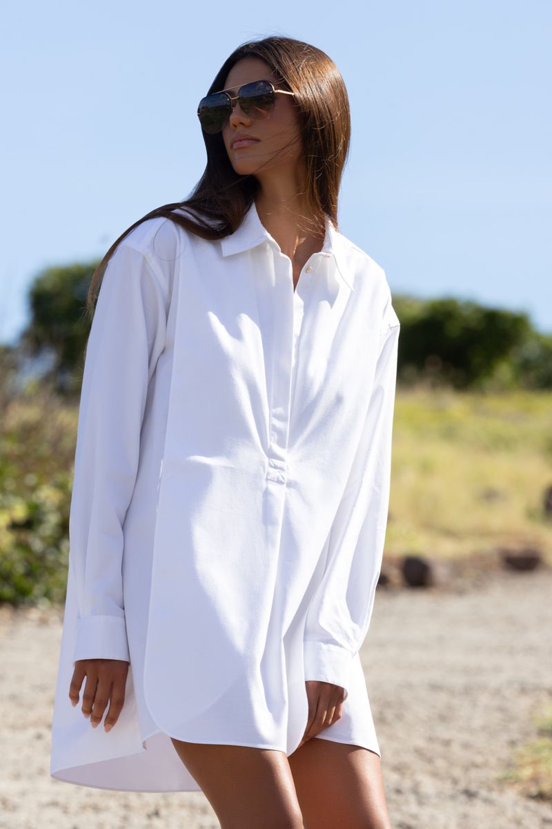 Woman wearing an oversized white collared shirt dress and dark sunglasses outdoors on a sandy coastal path