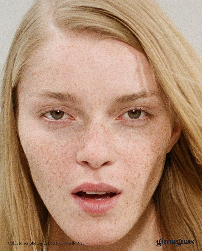 Close-up portrait of a fair-skinned woman with freckles and blonde hair, captured in soft, neutral lighting against a muted background.