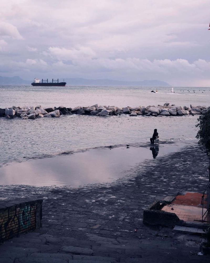 A solitary figure sits beside a reflective puddle on a stone waterfront overlooking a calm sea with a distant cargo ship.