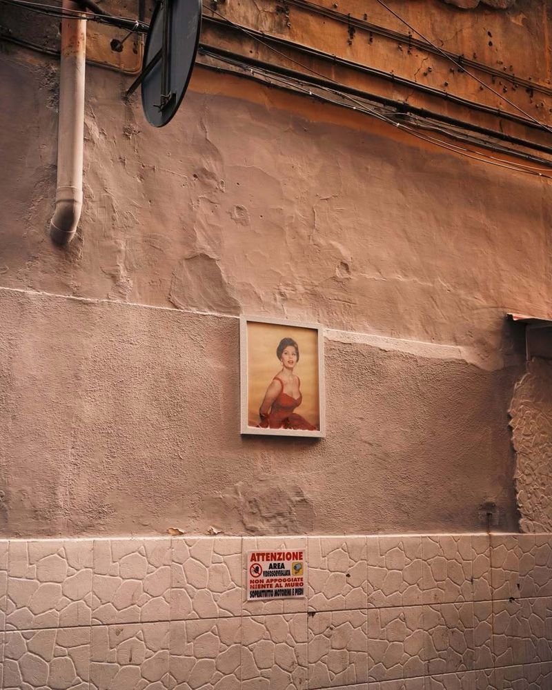 Framed portrait of Sophia Loren mounted on a weathered textured wall above decorative tiles in a Naples alley.