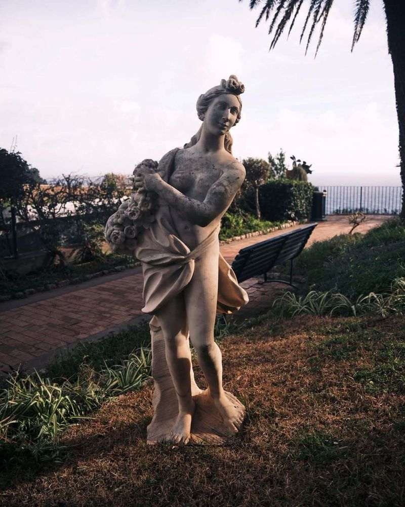 Classical stone statue of a draped woman standing on a grassy slope in a coastal garden with a park bench and paved walkway.
