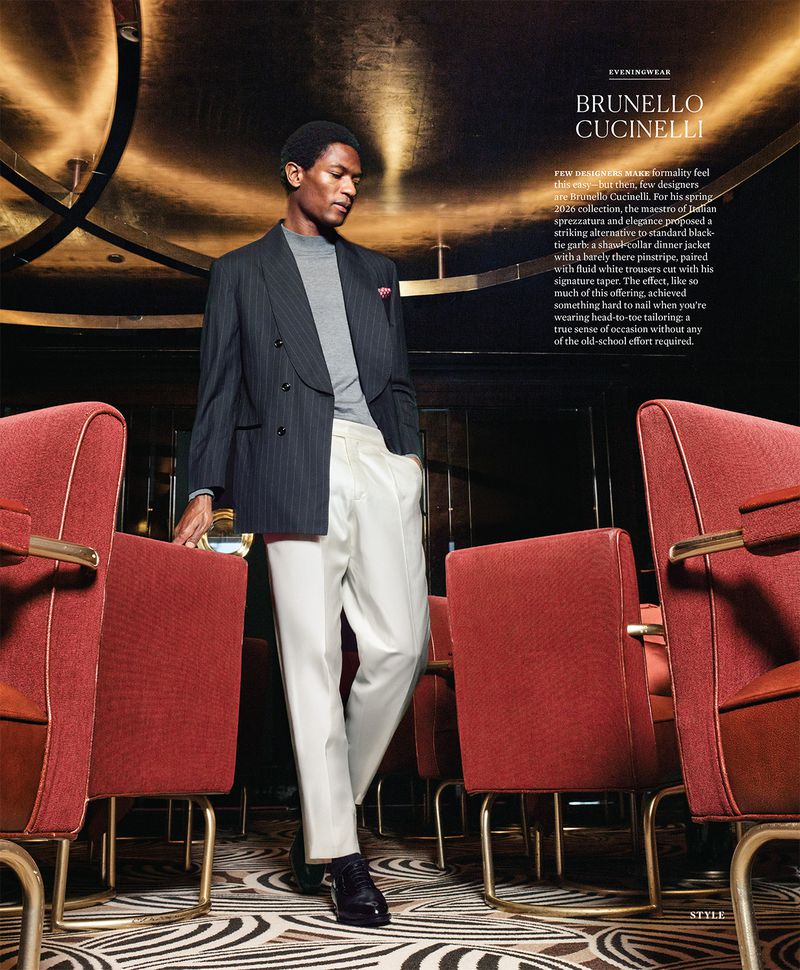 Hamid Onifadé wearing a pinstriped blazer and white trousers standing among red velvet chairs in a dimly lit lounge.