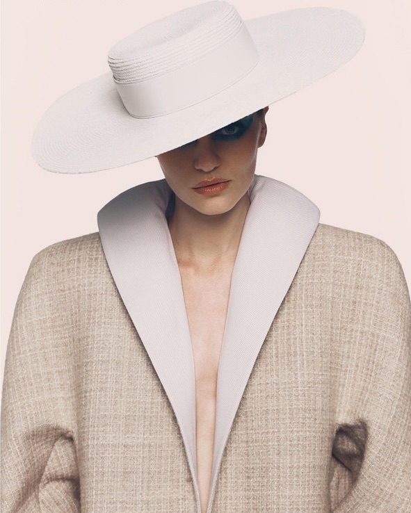 A woman with dramatic makeup wears a wide-brimmed hat and a large, open coat, posing against a soft pink backdrop in a fashion forward portrait.