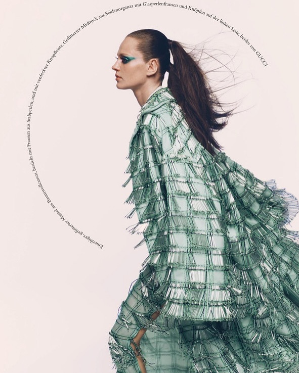 Editorial fashion shot of a woman with green eye makeup wearing a light green fringed jacket and skirt, posing indoors against a neutral backdrop.