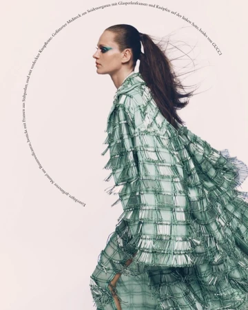 Editorial fashion shot of a woman with green eye makeup wearing a light green fringed jacket and skirt, posing indoors against a neutral backdrop.