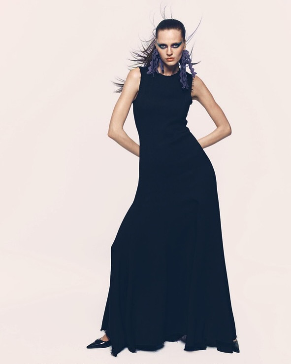 A fashionable adult female poses indoors wearing a full-length sleeveless black dress, earrings, and black shoes, in a studio setting.