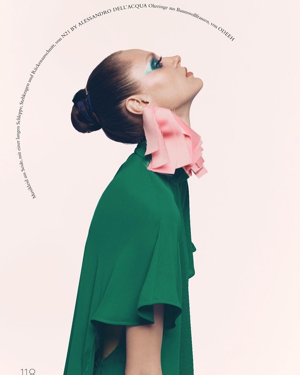 Fashion editorial shot of a woman in a green top with a pink ruffled collar, looking up against a pale pink background with text.