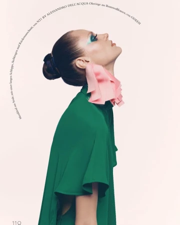Fashion editorial shot of a woman in a green top with a pink ruffled collar, looking up against a pale pink background with text.