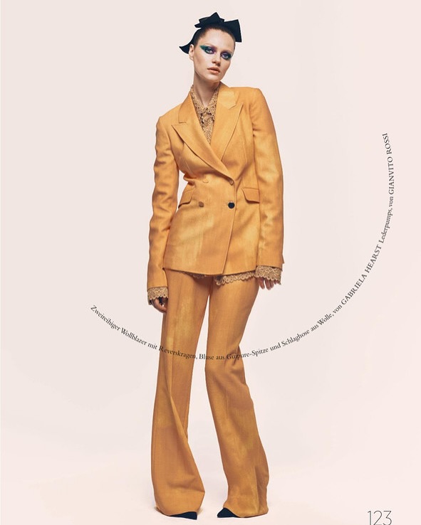 Fashion editorial shot of a woman in a yellow-gold blazer and matching wide-leg pants against a light background, modeling a high-fashion look.