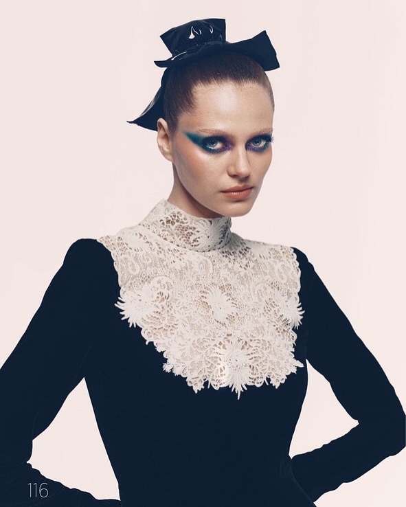 Fashion portrait of a female model wearing a navy dress with white lace collar, dramatic eye makeup, and a large black bow in her hair.
