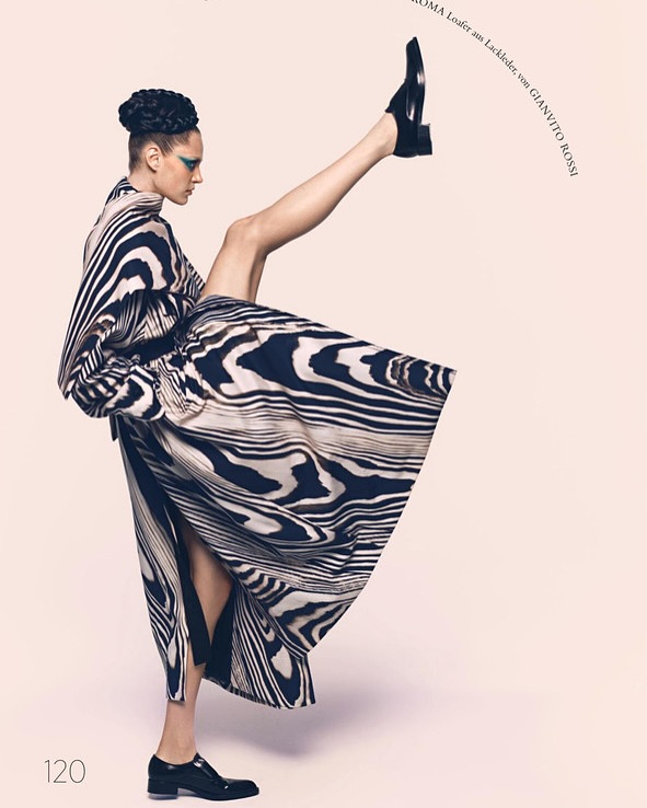 Fashion editorial shot of a woman in a patterned dress kicking one leg in the air, wearing patent leather shoes against a pink background.