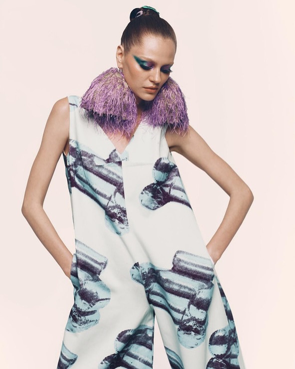 Elegant adult female fashion model poses indoors wearing patterned jumpsuit and purple feathered collar with dramatic makeup and styled hair.