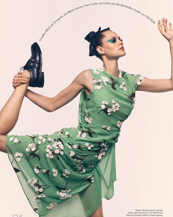 Fashion editorial shot of a woman in a green floral dress holding up a leather shoe in a contorted pose against a neutral background.