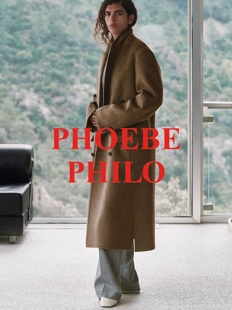 A woman in a tan coat and grey pants stands in front of a large window with a mountain view, showcasing a Phoebe Philo design.