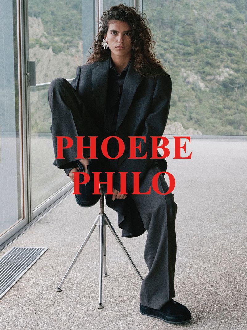A model poses on a stool in an oversized gray suit with a mountain view background and the text PHOEBE PHILO overlaid in red.