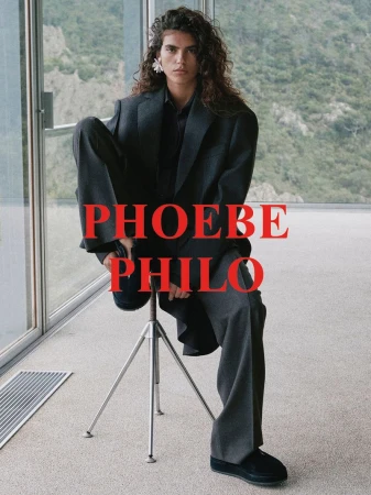 A model poses on a stool in an oversized gray suit with a mountain view background and the text PHOEBE PHILO overlaid in red.
