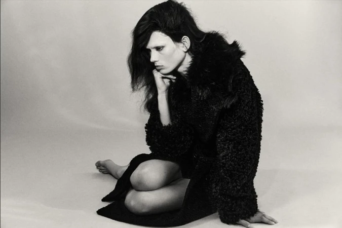 Model Wilda Rubin seated on a studio floor wearing an oversized black textured coat in a black and white editorial portrait.
