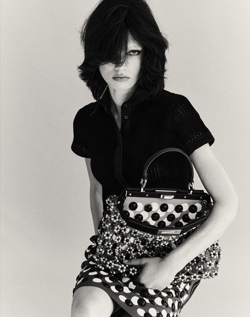 Model with dark layered hair wearing a black polo shirt holding a patterned handbag against a light background