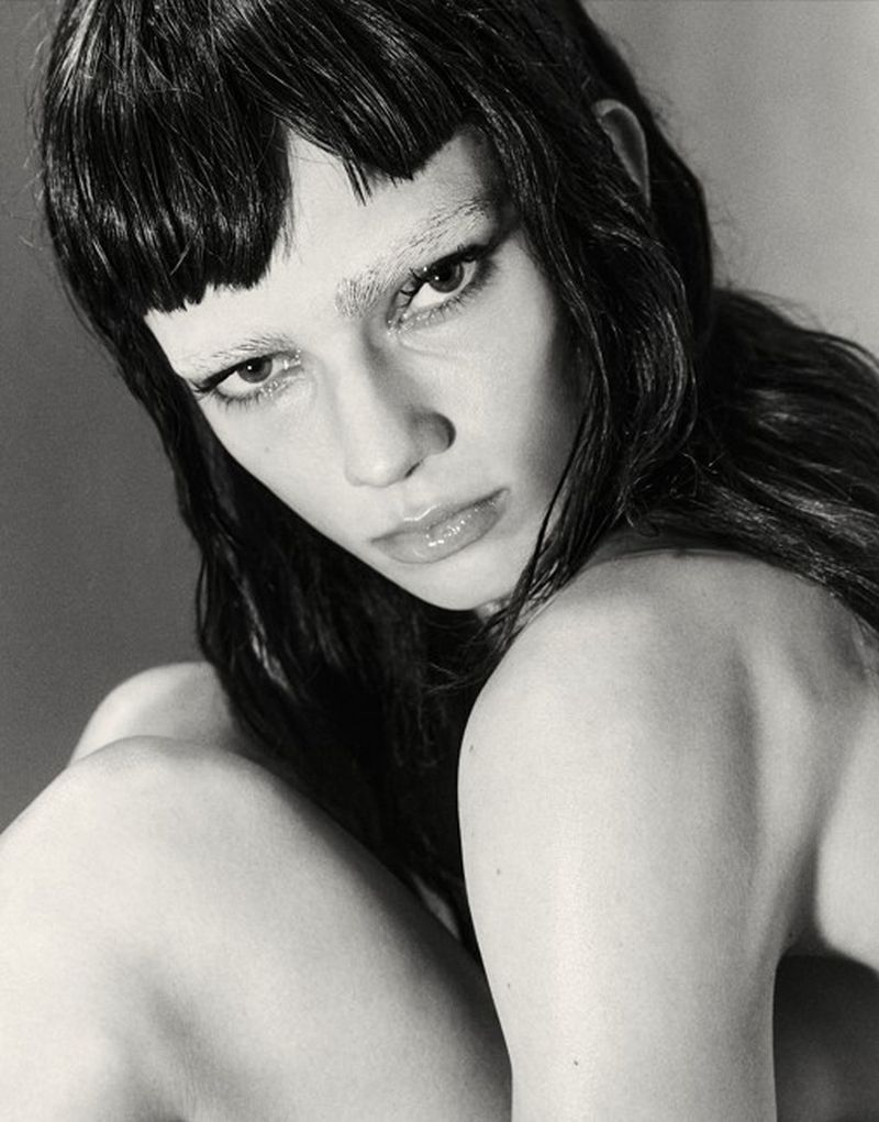 Close-up portrait of a woman with dark hair, blunt micro bangs, and bleached eyebrows against a neutral studio background.