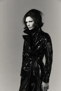 Model Wilda Rubin wearing a high-shine black patent leather belted trench coat in a monochrome studio portrait.