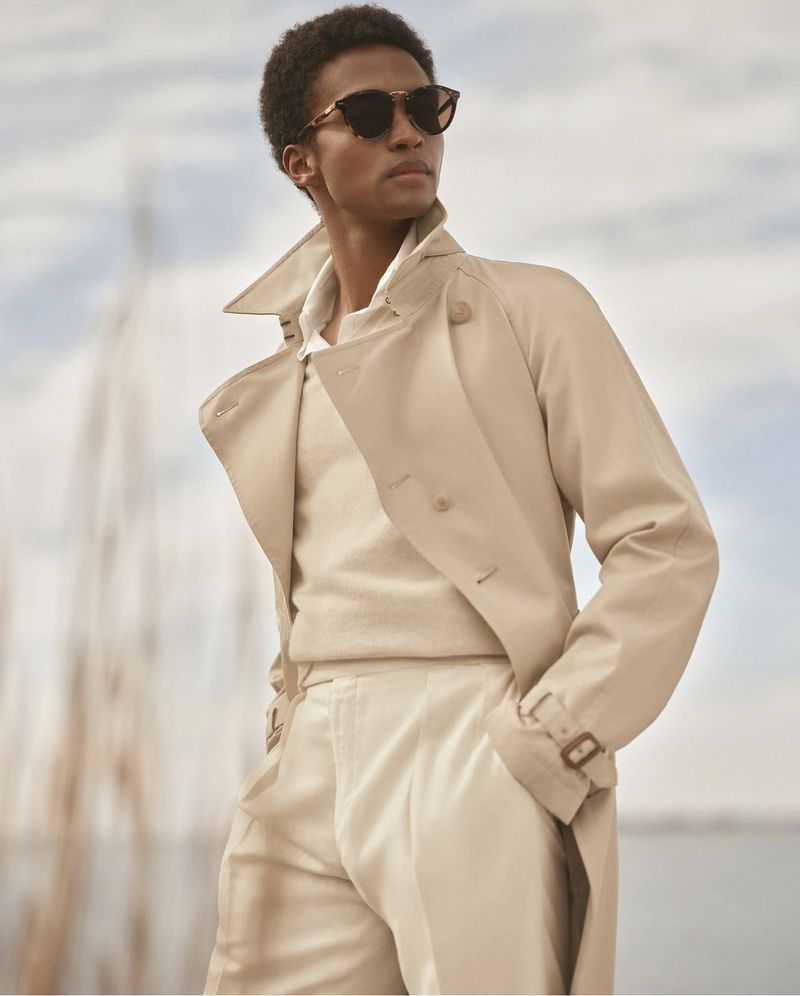 A young adult male poses in a beige trench coat and sunglasses with a blue sky background and reeds in the background, fashion style.