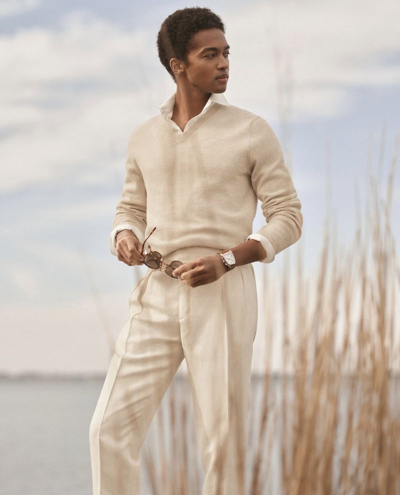 A stylish Black man in a beige sweater and pants holds sunglasses outdoors near a body of water and tall grasses under a cloudy sky.