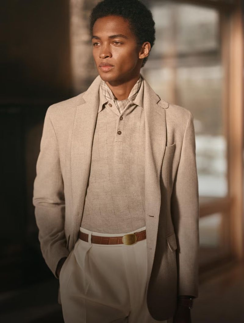 Portrait of a handsome man in a beige suit and white trousers, styled with a brown belt and collared shirt. Indoor fashion shot.