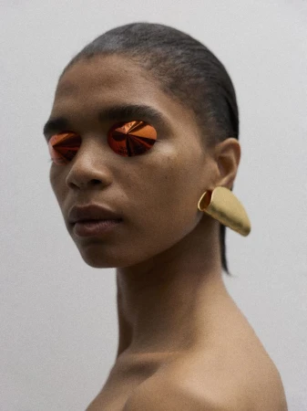 Model Raven Wallace wearing sculptural amber eye covers and a matte gold ear cuff against a neutral studio backdrop.