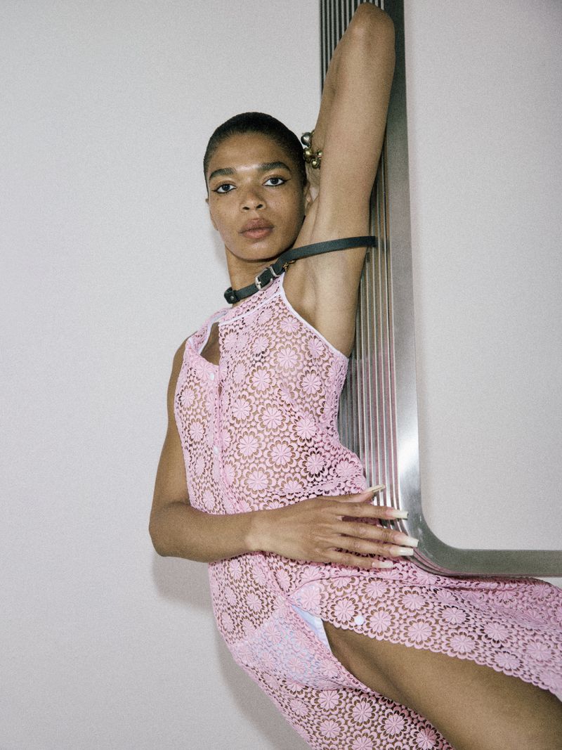 Model Raven Wallace posing in a sheer pink floral lace dress against a metallic fixture in a minimalist studio.