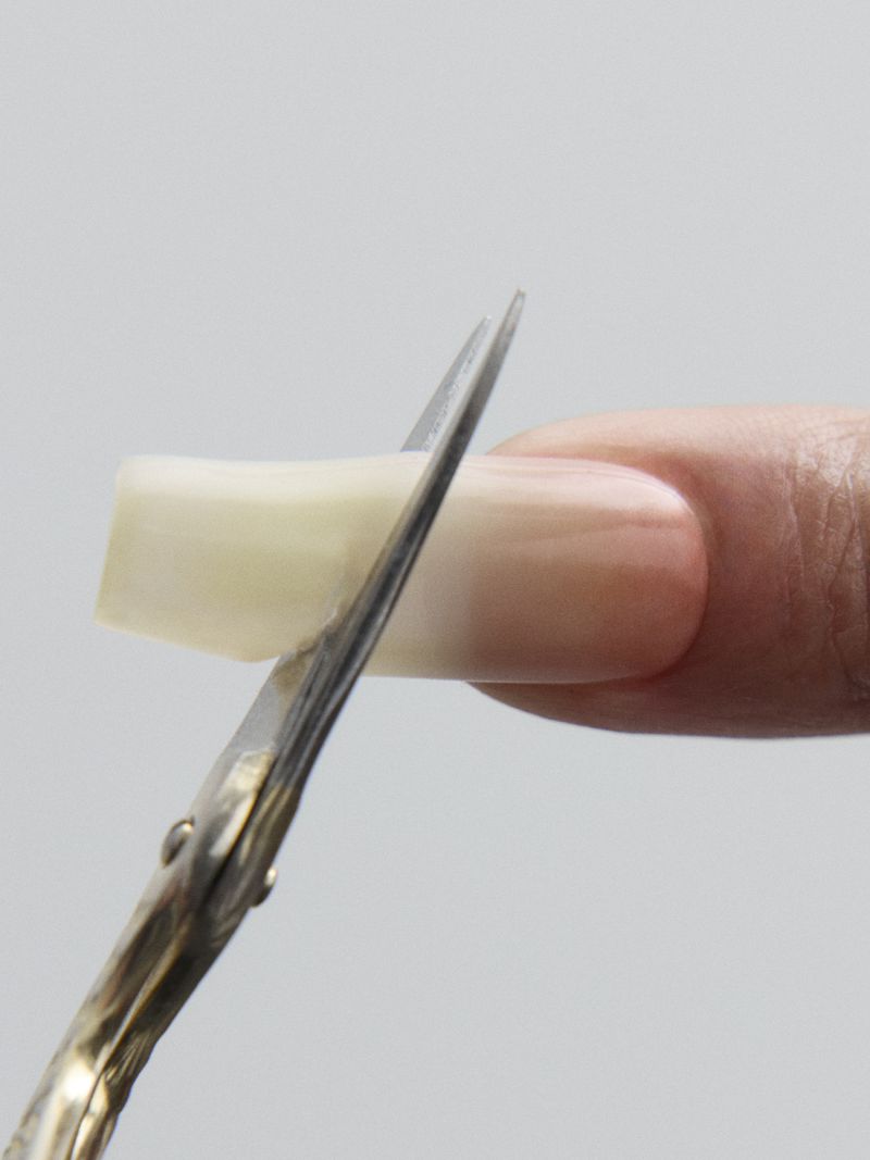 Close-up of ornate metallic manicure scissors trimming a long acrylic nail tip on a person's finger against a grey background.