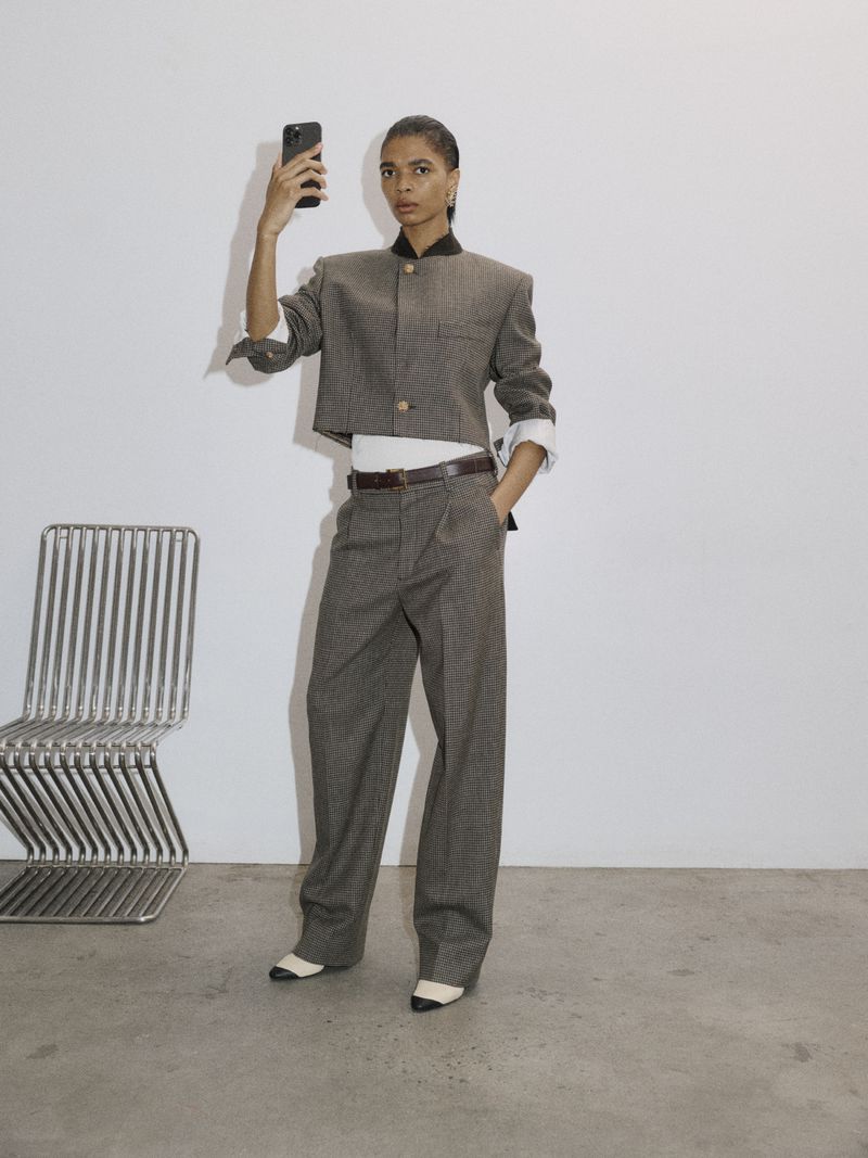 Model Raven Wallace posing with a smartphone in a cropped tweed jacket and wide-leg trousers beside a metal chair.